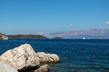 Kıyıda mavi sularda büyük kayalar, beyaz yelkenli yat ve ufukta dağlar, Yunanistan 'daki Lefkada adası. İyon Denizi 'ne yaz canlı doğa yolculuğu