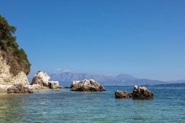 Uzak dağları ve parlak gökyüzü olan İyon Denizi 'nin çakıl taşlı kumsalında mavi sudaki manzara kayaları. Yunanistan 'daki Lefkada adasının doğası. Yaz tatilinde gidilecek yer