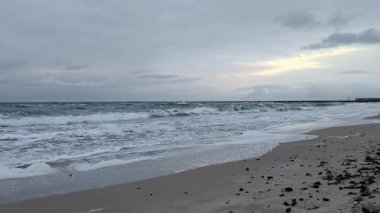 Soğuk akşam ışığında bulutlu gökyüzünün altında kumlu kıyılara çarpan dalgalarla dramatik deniz manzarası