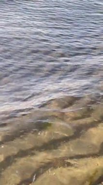 Güneş ışığı yansımaları ve pürüzsüz dalga hareketleriyle açık deniz suyu ve kumun dikey görüntüsü