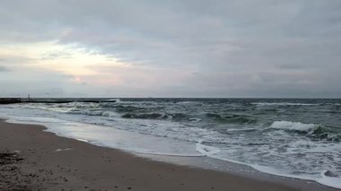 Bulutlu gökyüzü ve kumsalda dalgalarla dramatik bir akşam.