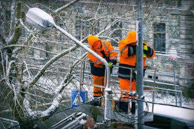 Turuncu üniformalı iki elektrikçi, elektrik direğinin tepesinde çalışıyor. Kışın şehir binalarının zemininde kablo hatları var..