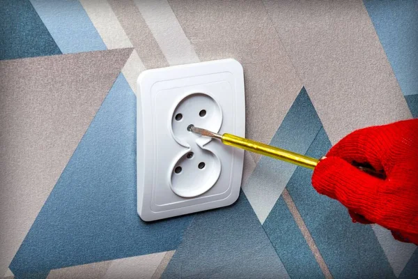 An electrician wearing red protective gloves uses a screwdriver to install a socket into the wall of the living room. Close-up of hands. Home master concept.