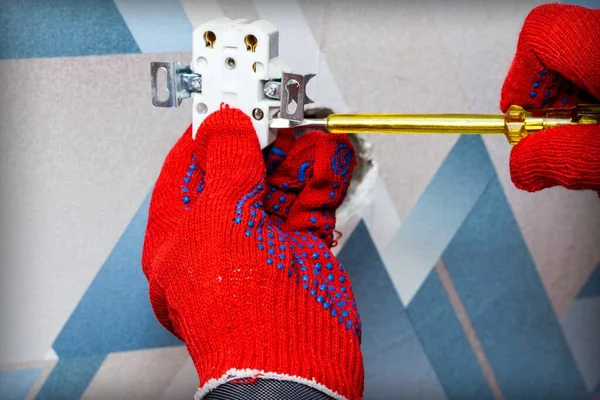 An electrician wearing red protective gloves uses a screwdriver to install a socket into the wall of the living room. Close-up of hands. Home master concept.
