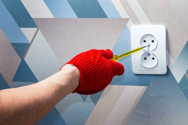 An electrician wearing red protective gloves uses a screwdriver to install a socket into the wall of the living room. Close-up of hands. Home master concept.