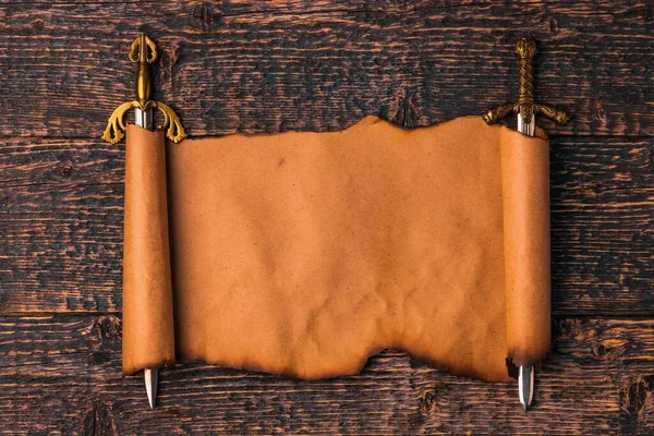 Rollo de papel viejo con bordes quemados estirados sobre las hojas de espadas de caballero sobre el fondo de la textura de madera vieja. — Foto de Stock