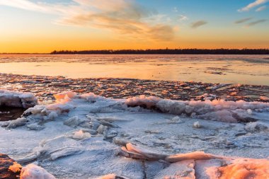 Gün batımında güneşin turuncu ışığıyla aydınlanan buzla kaplı bir nehrin manzarası. Kış manzarası