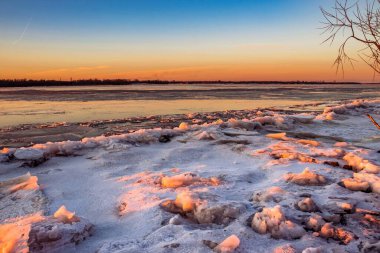 Gün batımında güneşin turuncu ışığıyla aydınlanan buzla kaplı bir nehrin manzarası. Kış manzarası