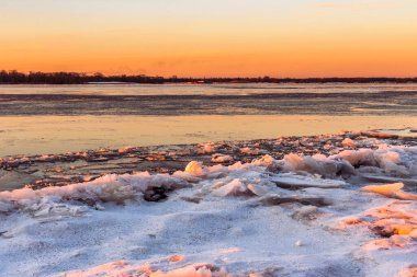 Gün batımında güneşin turuncu ışığıyla aydınlanan buzla kaplı bir nehrin manzarası. Kış manzarası