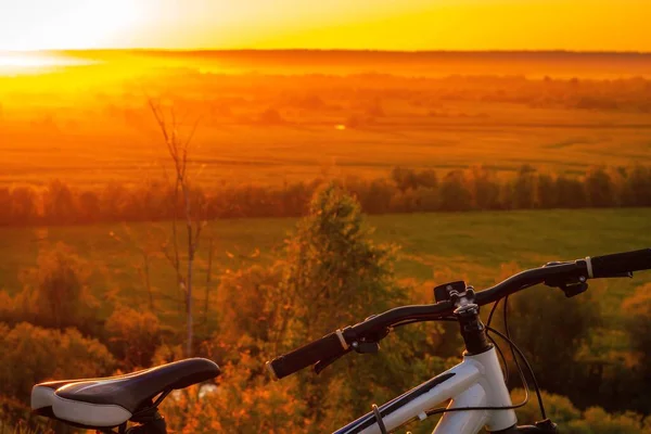 Bicicleta em uma colina contra o pano de fundo de um pôr do sol sobre um prado com grama verde e árvores. — Fotografia de Stock