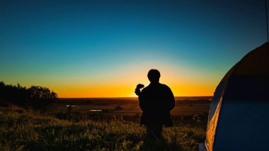 Çadırın yanında dürbünlü bir adamın silueti gün batımının arka planına karşı açık bir gökyüzünde çayırın üzerinde.