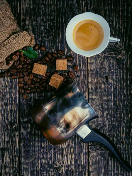 Cezve, a cup of coffee, a bag of spilled coffee beans and cane sugar lumps on an old wooden surface. Flat lay, vintage