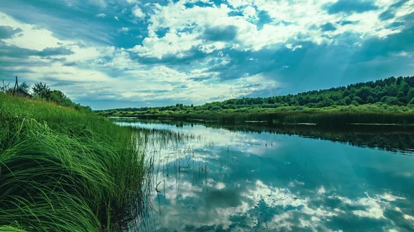 Die Wasseroberfläche des Flusses mit einer Reflexion des blauen Himmels mit Wolken, grüne Seggen entlang der Ufer. — Stockfoto