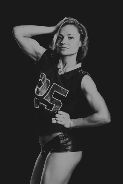 Sexig tjej med en perfekt figur klädd i en T-shirt amerikansk fotbollsspelare. Sexig athletic blondin poserar på en svart bakgrund. Muskulös tjej på en svart bakgrund. — Stockfoto