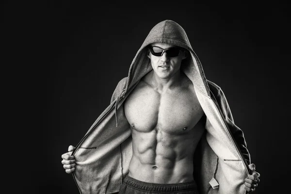 Homem musculado em uma jaqueta de esportes e óculos escuros em um fundo preto. Demonstração músculos do corpo durante a jaqueta esportiva. Sportswear e acessórios. Fotos para revistas e sites esportivos . — Fotografia de Stock