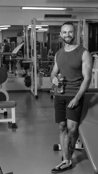 Een sterke man in de sportschool. Rust tussen de oefeningen. Knappe man drinkt water. — Stockfoto
