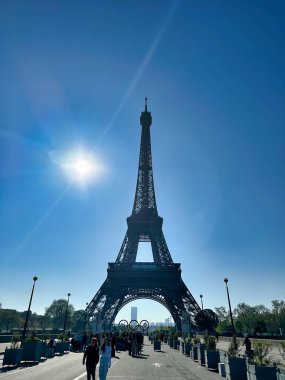 Güneşli bir günde Eyfel Kulesi 'nde insanlar aşağıda yürüyor ve arka planda Olimpiyat halkaları var..
