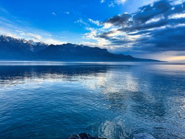 Sakin mavi sularla sakin göl manzarası, dağ siluetleri ve gün batımında dramatik bulutlu gökyüzü..