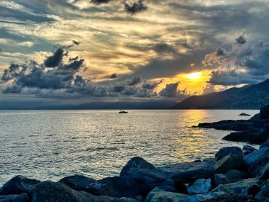 Deniz üzerinde dramatik sahil günbatımı parlayan bulutlar, ön planda kayalık kıyı ve ufukta kaybolan uzak dağlar..