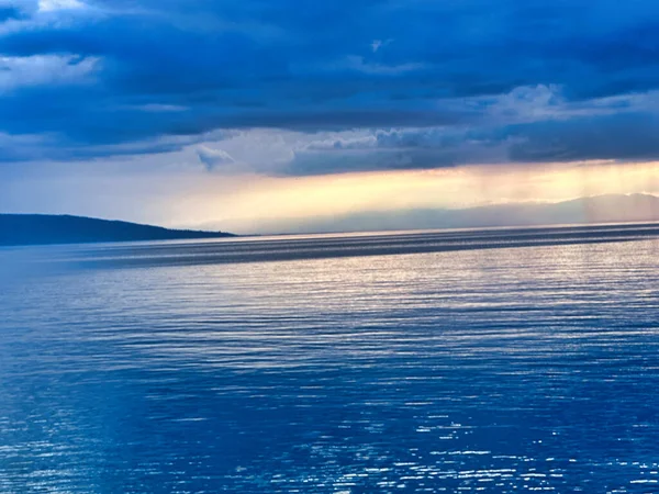 Sakin mavi sularla sakin göl manzarası, dağ siluetleri ve gün batımında dramatik bulutlu gökyüzü..