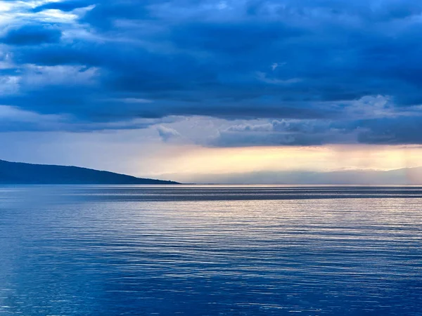 Sakin mavi sularla sakin göl manzarası, dağ siluetleri ve gün batımında dramatik bulutlu gökyüzü..