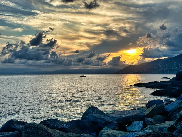 Deniz üzerinde dramatik sahil günbatımı parlayan bulutlar, ön planda kayalık kıyı ve ufukta kaybolan uzak dağlar..