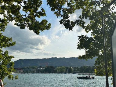 Yelkenliler dramatik bir gökyüzünün altında sakin bir gölde süzülür, uzak tepeler huzurlu sulara derinlik katar..