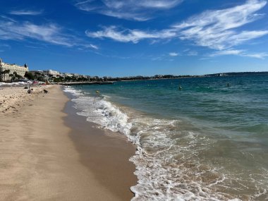 Turkuaz dalgalı güneşli kumlu sahil ve arka planda şehir silüeti..