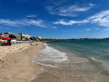 Turkuaz dalgalı güneşli kumlu sahil ve arka planda şehir silüeti..