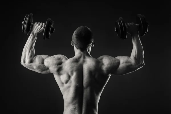 Homem muscular faz exercícios com halteres em um fundo escuro — Fotografia de Stock