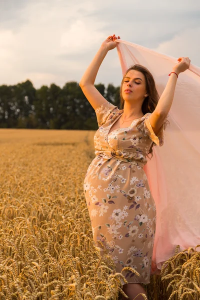 Terhes nő egy zsebkendőt, a kezében egy búza mezőben. Boldog asszony egy mezőben. Egy nő élvezi a születendő gyermek. Őszinte érzelmek és örömteli érzés. Gyönyörű vidéken. Búzamező vár betakarításra. — Stock Fotó