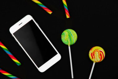 Siyah arka planda şekerlerin ve akıllı telefonun kavramsal tasarımı, sağlıksız yiyecekler, lolipoplar ve şekerler, en iyi görüntü.