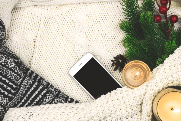 Modern winter cozy background, woolen and knitted sweater, fir branch with red berries and candles, blank smartphone