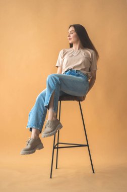 Stüdyoda bej arka planda sıradan giyinmiş genç bir kız. Eski sandalyede poz veren çekici kadın portresi. Yaşam tarzı kavramı 