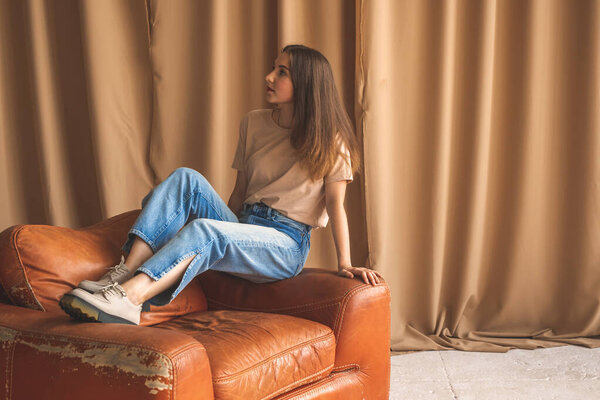 Teen girl in casual clothes posing in studio. Luxury old leather armchair and woman in denim jeans. Fashion and lifestyle background 