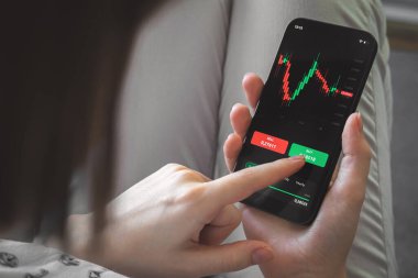 Cep telefonu uygulaması kullanılarak borsa ve kripto para birimi grafiklerinin analizi. Ekranda akıllı telefonu ve finansal grafikleri olan bir kadın. İş ve yatırım kavramı