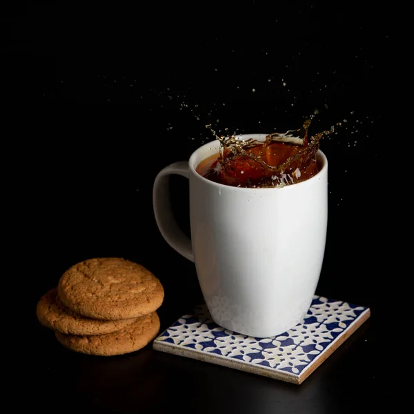 Glass with tea pouring with liquid with splashes and drops of water. Brewing tea concept. Cup pouring with water or tea with splashes on dark wooden background. Cup, teabag and cookies on table. — Stock Photo, Image