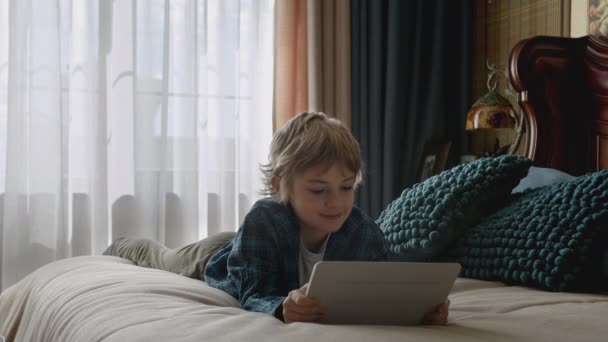 孩子们笑着躺在床上玩平板电脑 使用手持设备进行娱乐 通过互联网进行远距离交流 男孩有一个视频电话在家里 — 图库视频影像