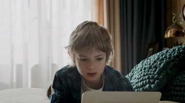 Çocuk gülümsüyor ve yatakta tabletle oyun oynuyor. Eğlence için el bilgisayarı kullanmak. İnternet üzerinden uzaktan iletişim. Çocuk evde video görüşmesi yapıyor..