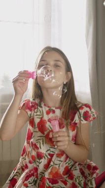 Gülümseyen çocuk oturma odasında eğleniyor, sabun köpüğü üflüyor. Mutlu küçük kız evde eğleniyor. Dikey çekim