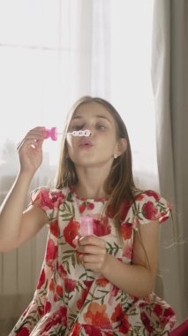 Gülümseyen çocuk oturma odasında eğleniyor, sabun köpüğü üflüyor. Mutlu küçük kız evde eğleniyor. Dikey çekim