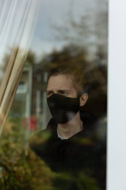 Evde yalnızlık çekerken pencereden dışarı bakan maskeli genç bir adam. Covid-19 Corona Virüsü Yalnızlık Konsepti