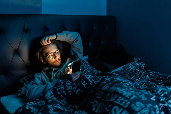 Sosyal medyaya bağımlı genç bir adam yatağa girmeden önce telefonunu kontrol ediyor. Bağımlılık, sosyal medya, karanlık bir kavram.
