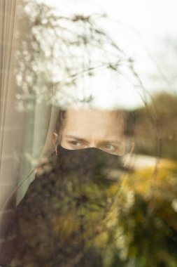 Evde yalnızlık çekerken pencereden dışarı bakan maskeli genç bir adam. Covid-19 Corona Virüsü Yalnızlık Konsepti