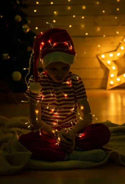 산타붉은 모자를 쓴 아이가 어두운 배경에 불빛으로 장식 된 복장을 하고 있습니다. 새해 마법이야. 크리스마스 컨셉 — 스톡 사진