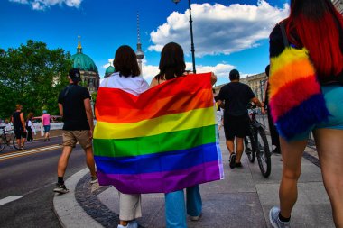 Berlin, Almanya - 26 Haziran 2021 - İki kadın Christopher Caddesi 'nde (CSD) gökkuşağı bayrağını paylaşıyor, arka planda Berlin TV kulesi ve Berlin Katedrali