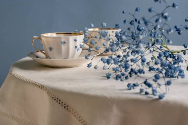 Porselen çay takımları ve mavi çiçekler beyaz masa örtüsü üzerinde.