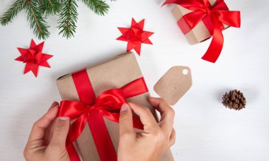 Yeni yıl kompozisyonu. Kadınların elleri hediye kutusuna fiyonk bağlıyor. Hediyeler, kırmızı kurdele ve arka planda ekolojik kağıttan bir zarf. Noel konsepti. Düz yatış, üst görünüm.
