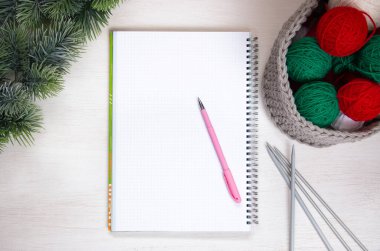 Gri bir sepetteki yeşil ve kırmızı iplik, notlar için bir defter, örgü iğneleri ve beyaz arka planda bir ladin dalı. Ev konforu ve Noel konsepti. Kadın ve erkeğin hobisi örgü örmektir..