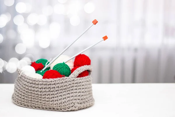 Green and red yarn in a knitted gray basket on a white table. Home comfort and christmas concept. Women\'s and men\'s hobby knitting.